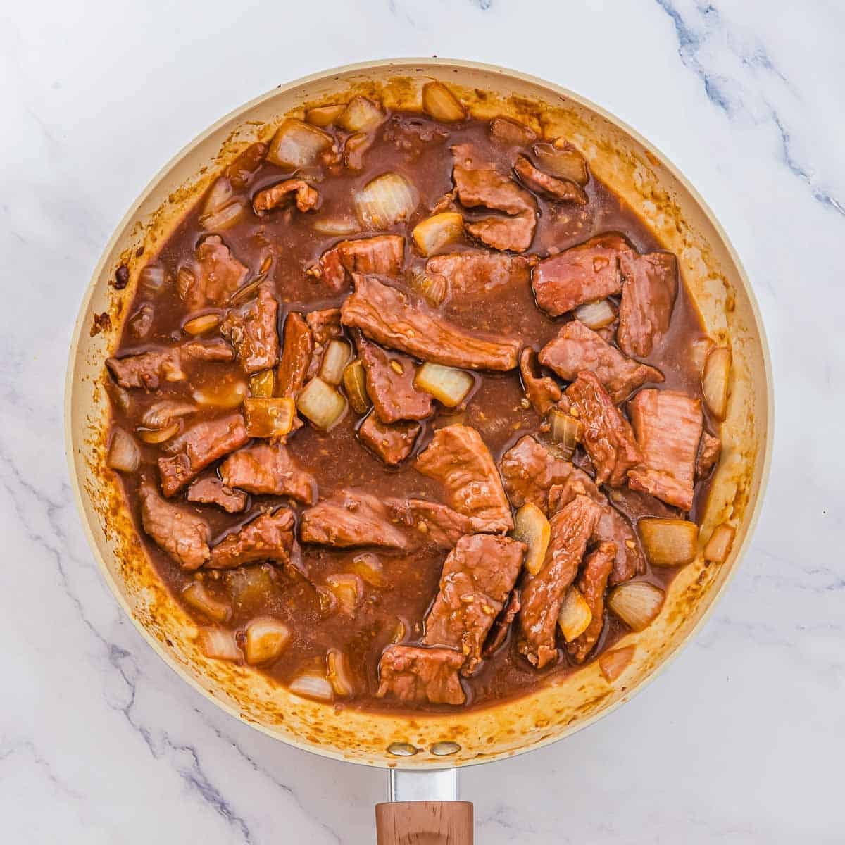 Beef and onion slices simmering in a brown savory sauce in a skillet.