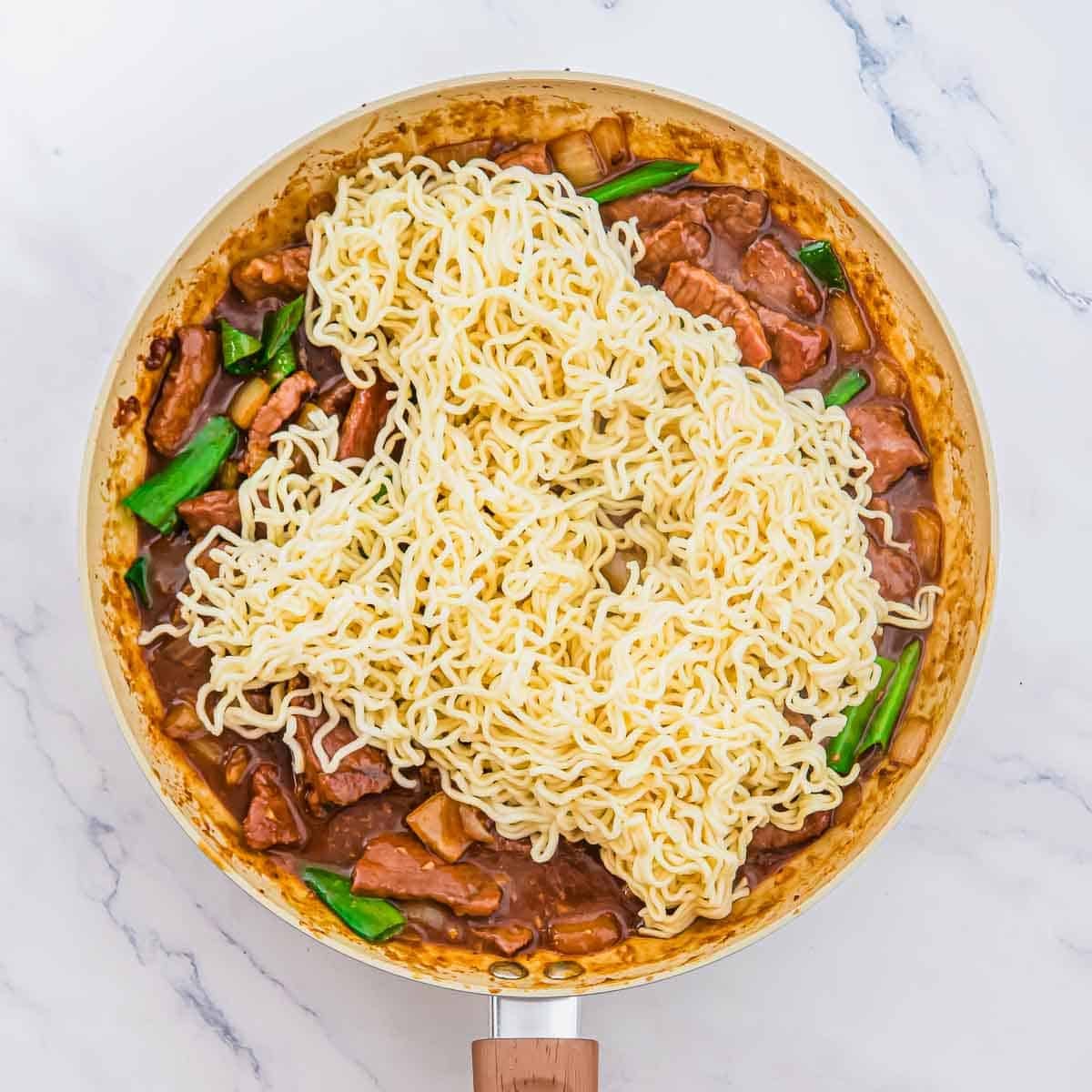 A large skillet containing stir-fried meat and green vegetables in brown sauce topped with cooked ramen noodles.