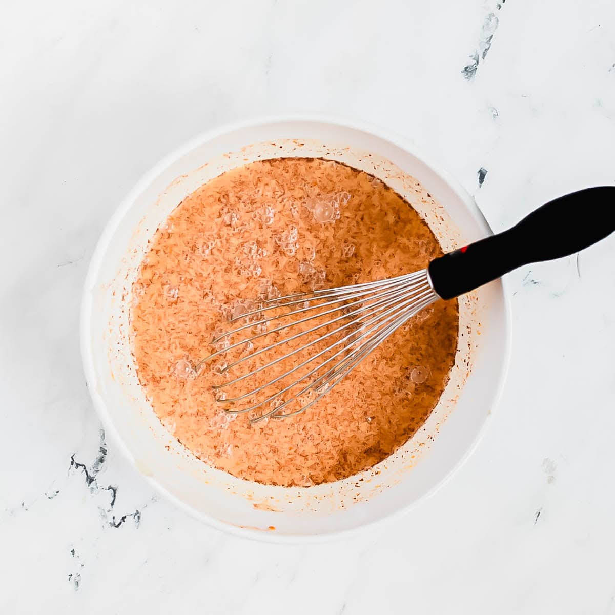 A whisk with a black handle rests in a white bowl filled with a pumpkin pudding mixture on a white marble surface.