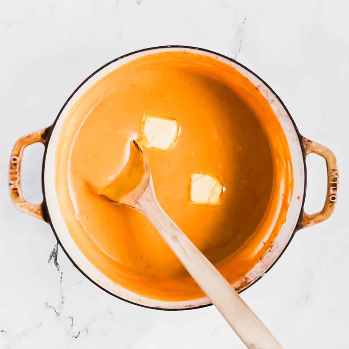 A pot of creamy orange pudding with two pats of butter melting on top, stirred with a wooden spoon, on a white marble surface.