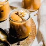 A small glass jar filled with creamy pumpkin pudding, topped with whipped cream and chopped nuts, sits on a plate next to a spoon.