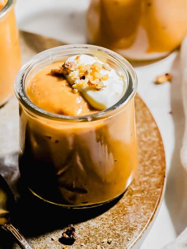 A small glass jar filled with creamy pumpkin pudding, topped with whipped cream and chopped nuts, sits on a plate next to a spoon.