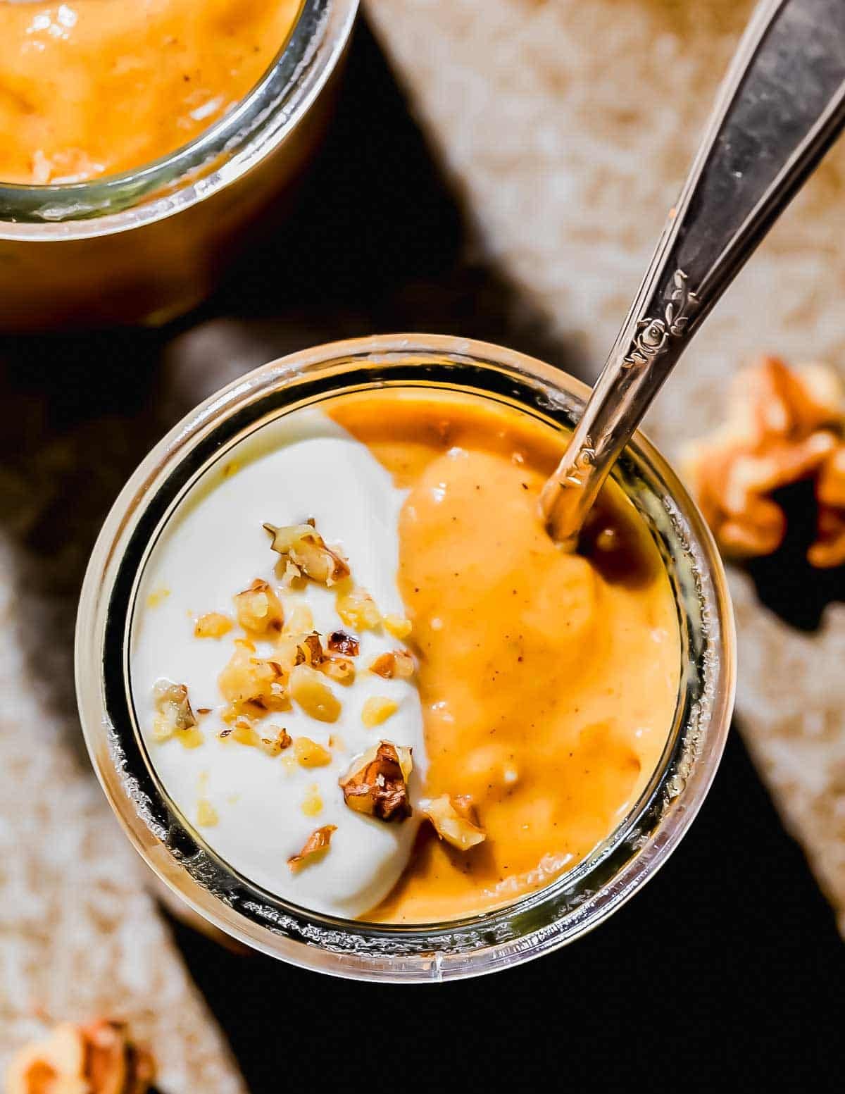 A glass jar filled with creamy pumpkin pudding, half topped with whipped cream and chopped walnuts, with a spoon inside.